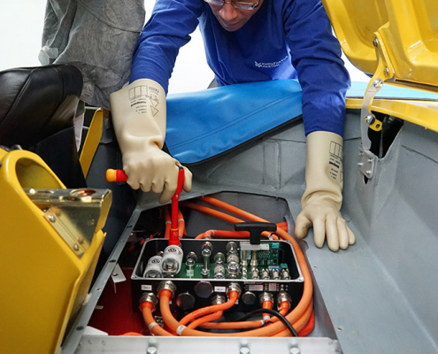 DSC05709_web Unter dem Verdeck-Raum einer gelben Corvette C1 sieht man elektronische Komponenten. Ein Ladegerät sowie eine Hochvolt-Stromverteilerbox. Die Stromverteilerbox ist offen und wird gerade von einer Person mit Schutzhandschuhen angeschlossen.