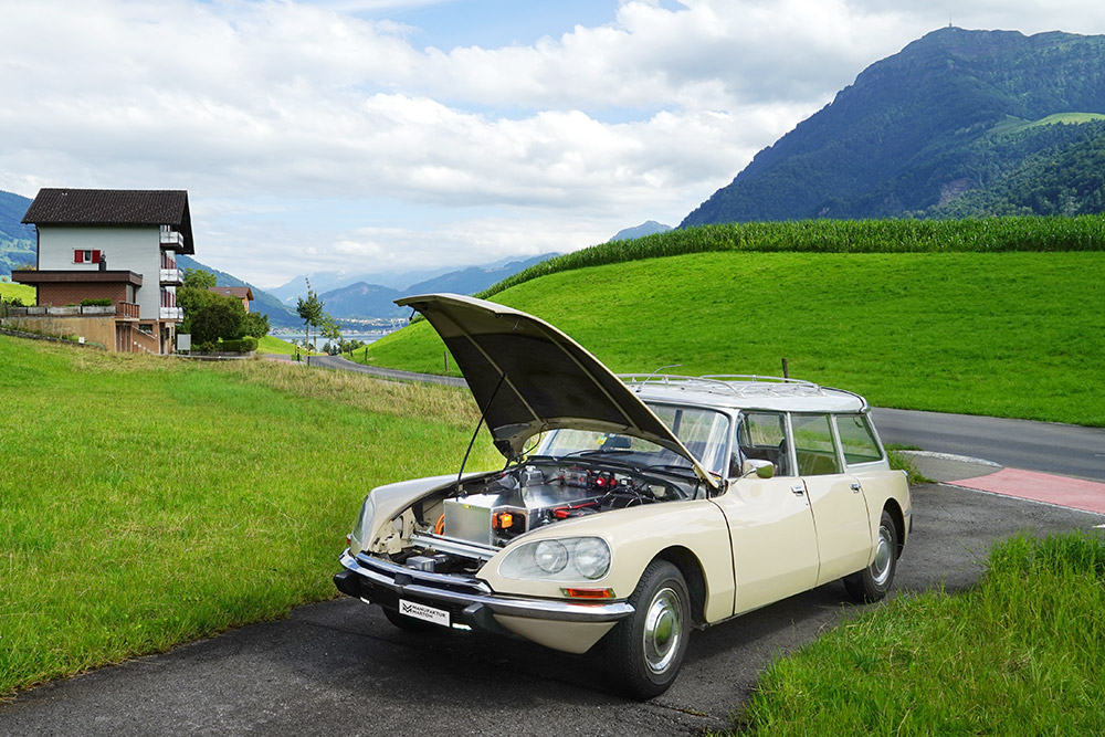 citroends23break_galerie28 Beige Citroen DS Break steht mit offener Motorhaube auf einem Einfahrtssträsschen im Grünen. Statt des Motors sieht man eine Batteriekiste. Im Hintergrund die Rigi.