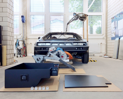 DJI_0068_deLorean_manufakturmarton_galerie5 Im Vordergrund die Batteriekiste und deren Deckel. Dahinter die Motorenhalterung und der Motor. Im Hintergrund das Fahrzeug von vorne, eine Flügeltüre geöffnet.