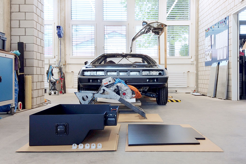 DJI_0068_deLorean_manufakturmarton_galerie5 Im Vordergrund die Batteriekiste und deren Deckel. Dahinter die Motorenhalterung und der Motor. Im Hintergrund das Fahrzeug von vorne, eine Flügeltüre geöffnet.