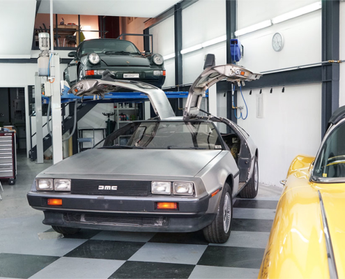 DSC06375_deLorean_manufakturmarton_gallerie2 DeLorean DMC-12 in einer Werkstatt. Von vorne, leicht seitlich. Die Flügeltüren sind offen. Im Hintergrund ein Porsche 911 auf einem Fahrzeuglift. Im Vordergrund ein Teil einer gelben Corvette C1.