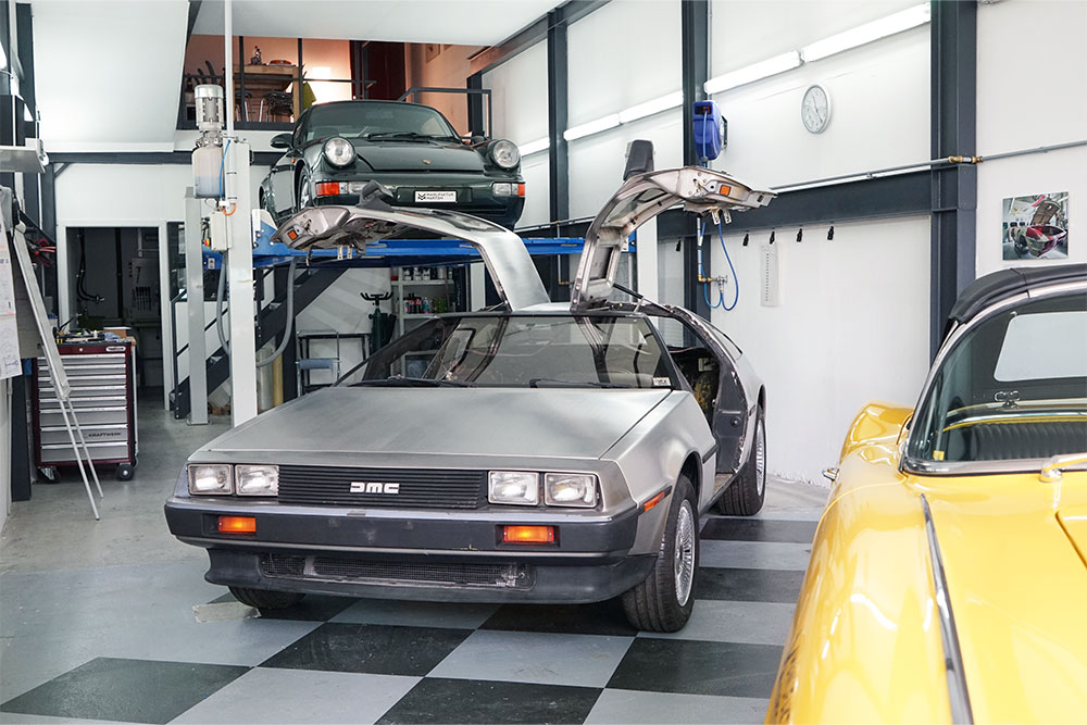 DSC06375_deLorean_manufakturmarton_gallerie2 DeLorean DMC-12 in einer Werkstatt. Von vorne, leicht seitlich. Die Flügeltüren sind offen. Im Hintergrund ein Porsche 911 auf einem Fahrzeuglift. Im Vordergrund ein Teil einer gelben Corvette C1.