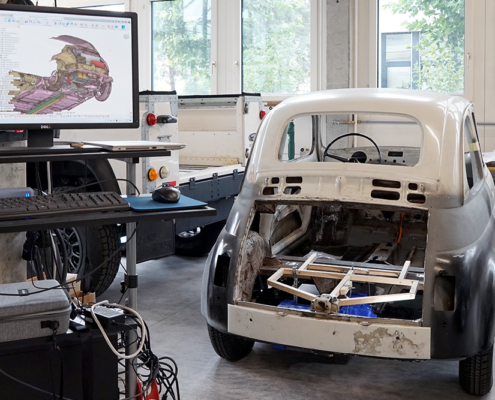 Ein Fiat Nuova 500 im Rohbau. Links davon ein Bildschirm, auf dem ein 3D-Scan des Fahrzeugs zu sehen ist.
