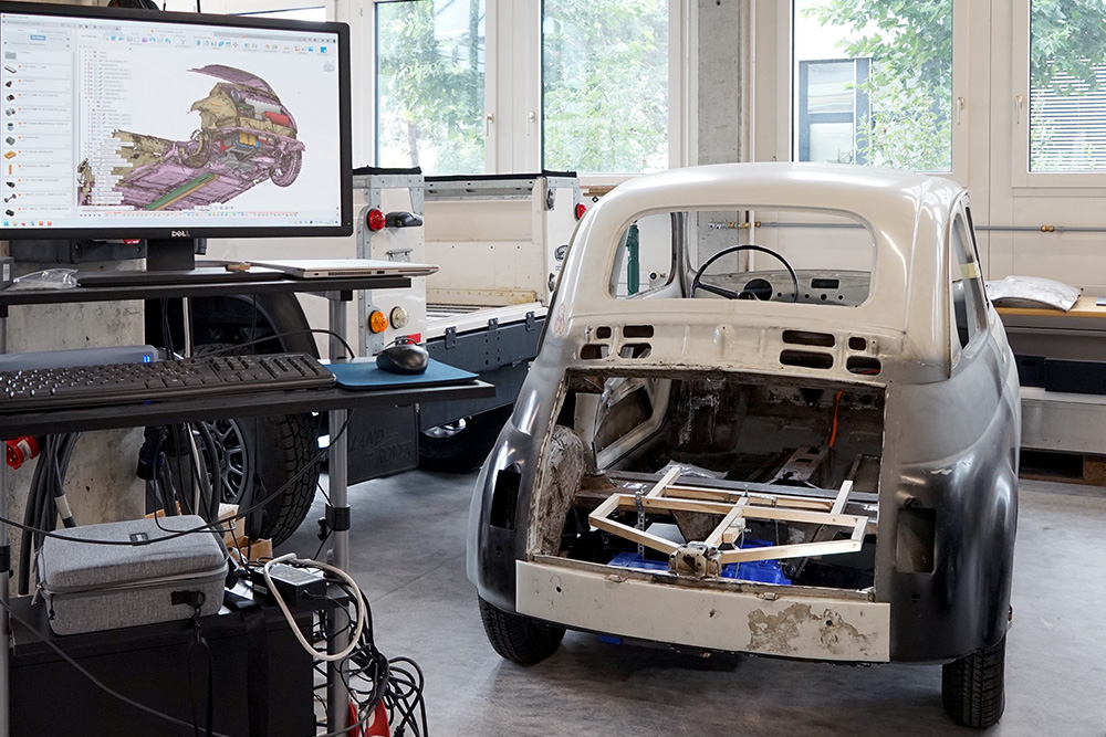 Ein Fiat Nuova 500 im Rohbau. Links davon ein Bildschirm, auf dem ein 3D-Scan des Fahrzeugs zu sehen ist.