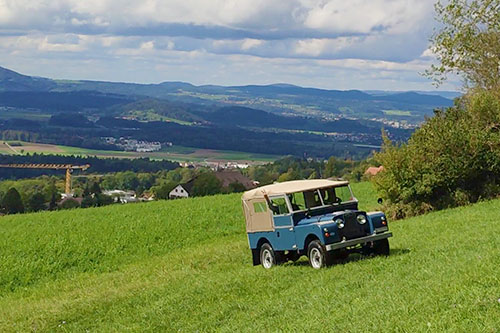 VID_20230919_140331_sabi3_-1-(00.00.30.08)_beitragsbild Ein blauer Landrover Series I mit beigem Verdeck fährt in einer hügeligen Landschaft eine Wiese hoch.