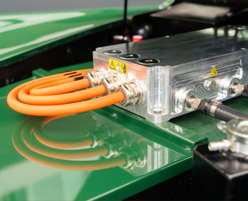 Blick in den Motorenraum des Jaguars. Man sieht den Deckel der Batteriekiste, in Wagenfarbe lackiert (grün), sowie den Umrichter, von welchem aus drei dicke, orange Hochvolt-Kabel im Bogen nach hinten gezogen werden.