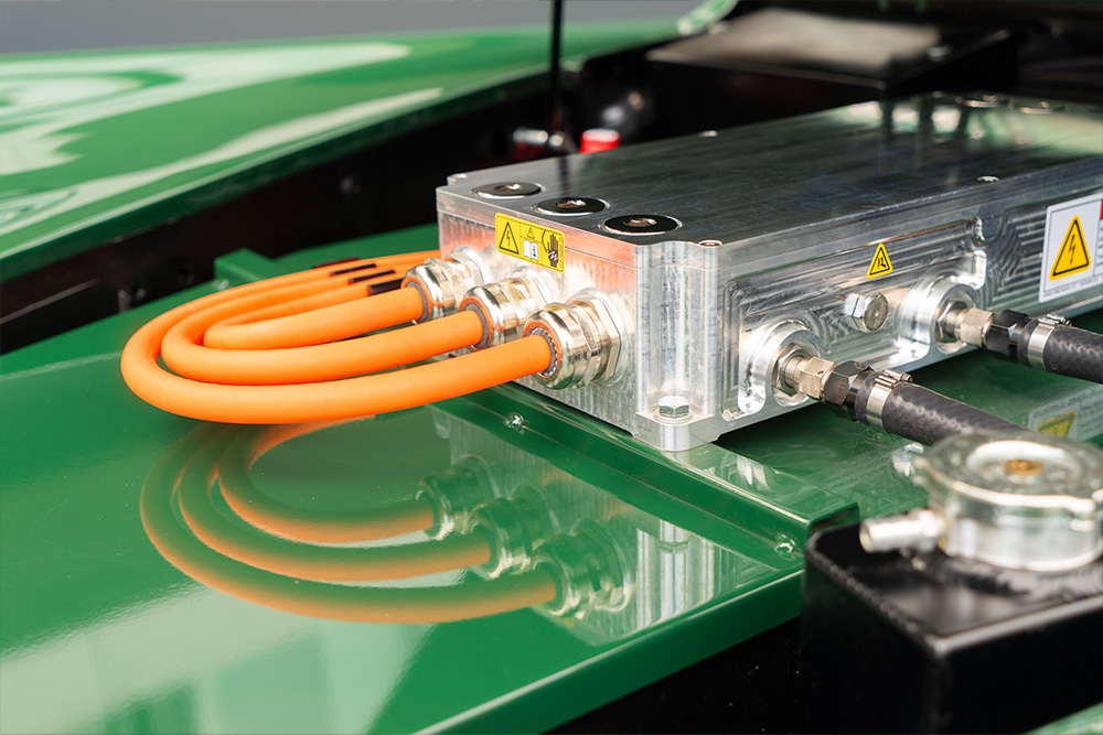 Blick in den Motorenraum des Jaguars. Man sieht den Deckel der Batteriekiste, in Wagenfarbe lackiert (grün), sowie den Umrichter, von welchem aus drei dicke, orange Hochvolt-Kabel im Bogen nach hinten gezogen werden.