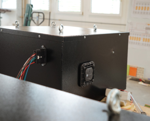 eLandy_manufakturmarton_galerie_03298 Blick auf die vordere Batteriekiste des eLandys, vor deren Einbau. Die Kiste ist schwarz. Man sieht den Niedervolt-Stecker mit diversen Kabeln, sowie ein Ventil zur Entgasung.