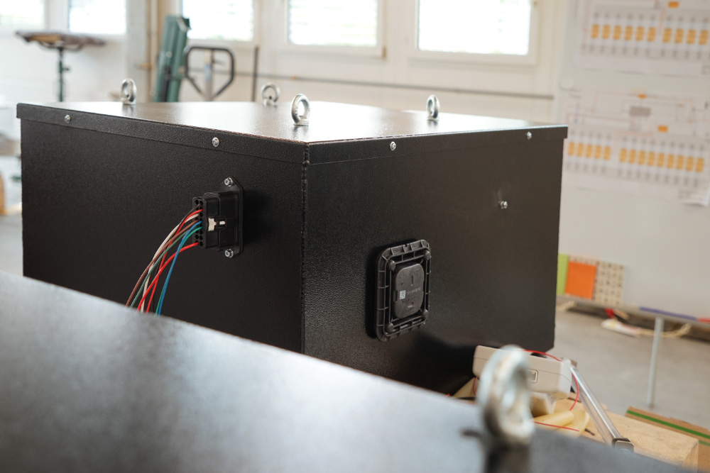 eLandy_manufakturmarton_galerie_03298 Blick auf die vordere Batteriekiste des eLandys, vor deren Einbau. Die Kiste ist schwarz. Man sieht den Niedervolt-Stecker mit diversen Kabeln, sowie ein Ventil zur Entgasung.