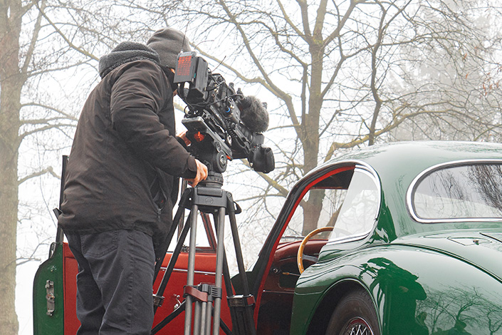 jaguarXK150_manufakturmarton_blog_047781 Ein Kameramann von hinten. Er steht neben einem Jaguar XK 150 mit offener Türe, und filmt durch diese das Interieur des Fahrzeugs. Im Hintergrund leicht vom Nebel verhüllte spätwinterliche Bäume.