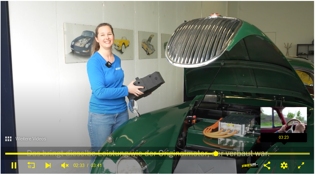 Screenshot aus Video von Streetlife Ausschnitt aus einem Video. Ein Jaguar XK 150 mit offener Fronthaube, im ehemaligen Motorenraum sieht man Batterie und Umrichter. Daneben Silvia Marton in blauem Shirt, in der Hand ein Dummy des im Fahrzeug verbauten Elektromotors.
