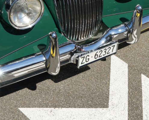 Jaguar XK 150 mit ZG Nummer Die Front eines grünen Jaguar XK 150, mit Fokus auf dem Nummernschild. Man sieht die verchromte Stossstange, einen der runden Scheinwerfer und einen Teil des Kühlergrills. Auf dem Boden sieht man einen Teil des Logos der Manufaktur Marton, ein stilisiertes Doppel-M.