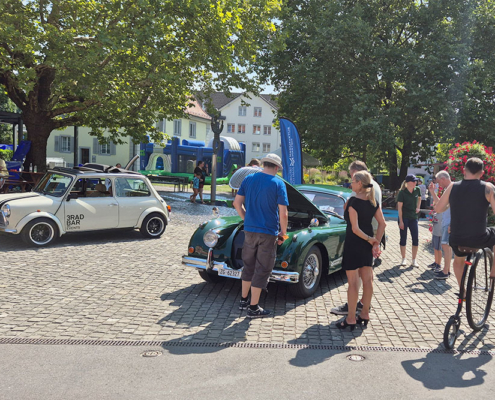 20240901_134455_blog Leute stehen um einen grünen Jaguar XK150 mit offener Fronthaube. Jemand fährt auf einem alten Hochrad vorbei.