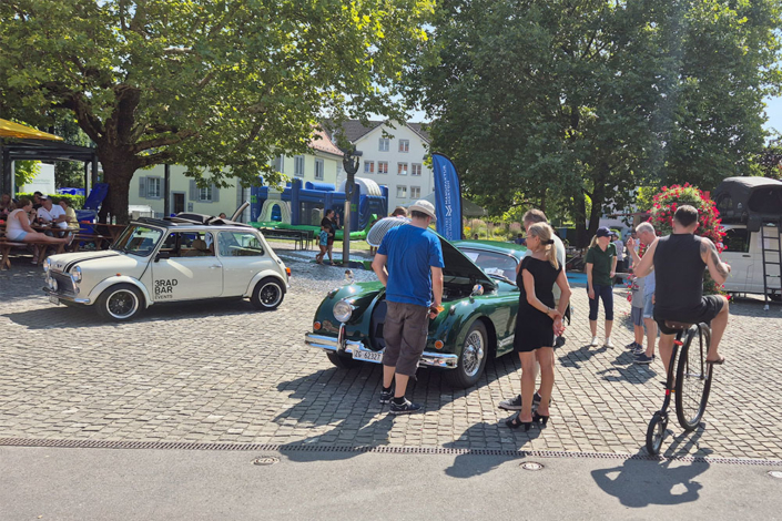 20240901_134455_blog Leute stehen um einen grünen Jaguar XK150 mit offener Fronthaube. Jemand fährt auf einem alten Hochrad vorbei.