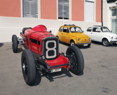 IMG_20240901_135748_blog Links ein roter Nash Twin Ignition Eight in offener Bauweise. Sein Kühlergrill wird von einer 8 geziert. Rechts davon zwei Fiat Nuova 500, einer in gelb, einer in weiss.