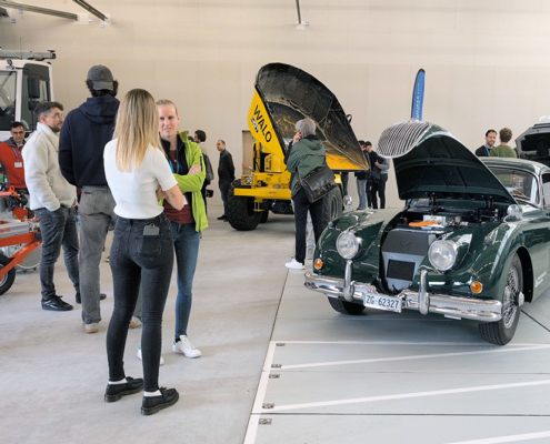 Galerie_1748436351285 Verschiedene Fahrzeuge, unter anderem ein Muldenfahrzeug, ein Mäher sowie ein Jaguar XK 150 stehen in einem Hangar. Diverse Menschen betrachten und besprechen die Fahrzeuge