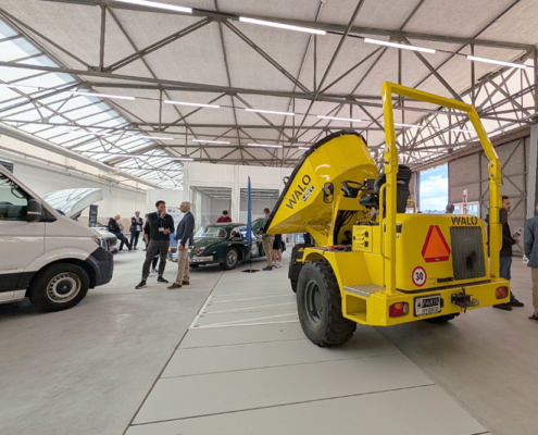 Galerie_1748436950825 Verschiedene Fahrzeuge, unter anderem ein Muldenfahrzeug, ein Lieferwagen sowie ein Jaguar XK 150 stehen in einem Hangar. Diverse Menschen betrachten und besprechen die Fahrzeuge