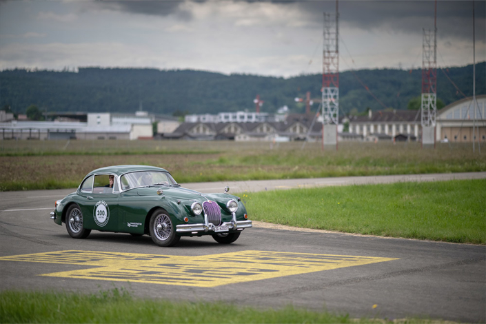 Jaguar auf Flugfeld Ein grüner Jaguar XK 150 fährt über eine Flughafen-Fahrbahn. Gelbe Marierungen auf der Fahrbahn, im Hintergrund Baumarkierungen. Auf der Seite des Jaguars ist eine Beschriftung angebracht, die darauf hinweist, dass es sich um ein Elektroauto handelt.