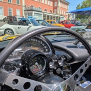 Blick über das Lenkrad einer Corvette C1. Die Armaturen verraten, dass es sich hier um ein Fahrzeug handelt, dass zum Elektroauto umgebaut wurde. Denn an Stelle des Tourenzählers zeigt die klassische Rundanzeige die Restreichweite an. Im Hintergrund sieht man diverse Fahrzeuge. Rechts eine Reihe mit diversen Fahrzeugen. Zwei Porsche 911 in weiss und grau, zwei Fiat Nuova 500 in gelb und rot sind gut erkennbar, dahinter lassen sich zwei Alfa Romeo GTV in rot und blau sowie eine Charleston-Ente erahnen.