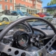 Blick über das Lenkrad einer Corvette C1. Die Armaturen verraten, dass es sich hier um ein Fahrzeug handelt, dass zum Elektroauto umgebaut wurde. Denn an Stelle des Tourenzählers zeigt die klassische Rundanzeige die Restreichweite an. Im Hintergrund sieht man diverse Fahrzeuge. Rechts eine Reihe mit diversen Fahrzeugen. Zwei Porsche 911 in weiss und grau, zwei Fiat Nuova 500 in gelb und rot sind gut erkennbar, dahinter lassen sich zwei Alfa Romeo GTV in rot und blau sowie eine Charleston-Ente erahnen.