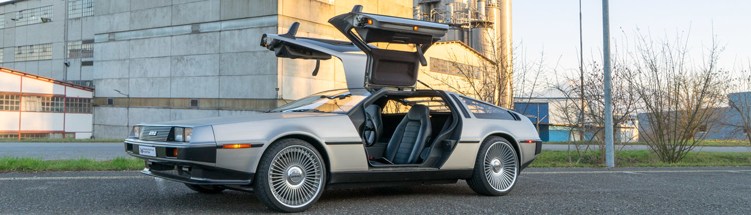 DeLorean_manufakturmarton_1500x430_projekt DeLorean DMC mit offenen Flügeltüren und massgeschneiderten Felgen vor Industriegebäude.