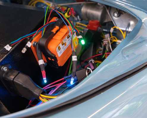 DSC08917_manufakturmarton_webgallerie Der Frontraum eines Fahrzeugs. Es sind diverse Kabel sichtbar, an einigen sind LEDs in verschiedenen Farben verbaut. An einer schwarzen Box, der Batteriekiste, ist ein oranger Trennschalter verbaut.