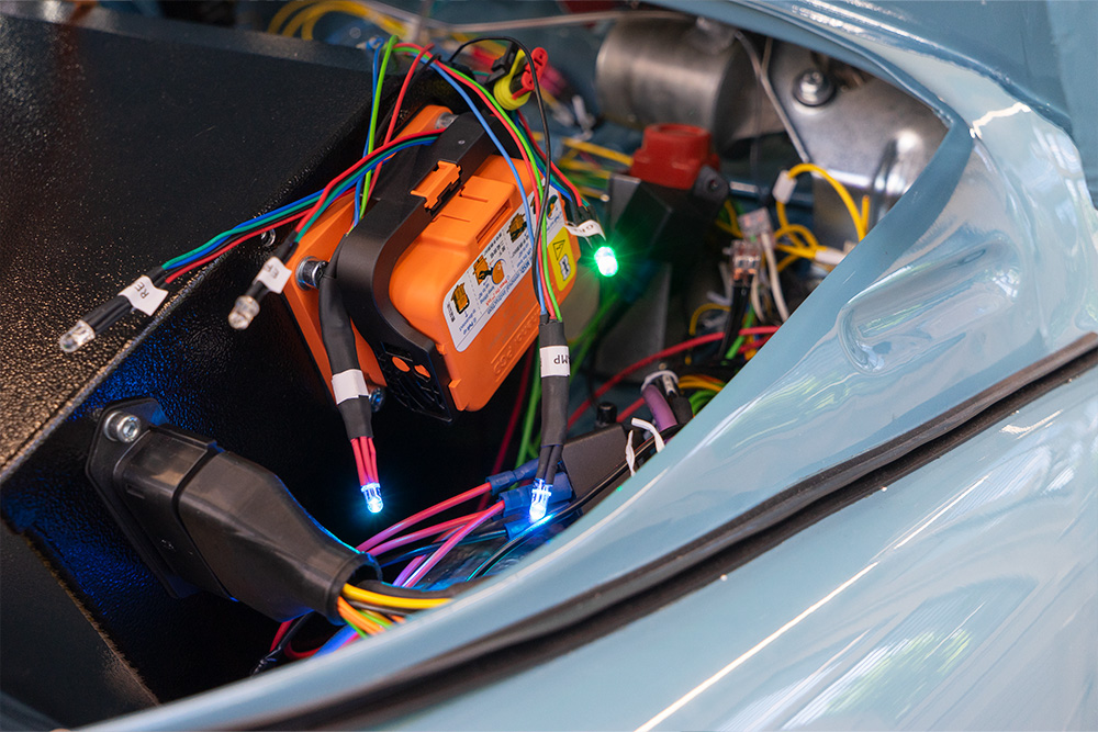 DSC08917_manufakturmarton_webgallerie Der Frontraum eines Fahrzeugs. Es sind diverse Kabel sichtbar, an einigen sind LEDs in verschiedenen Farben verbaut. An einer schwarzen Box, der Batteriekiste, ist ein oranger Trennschalter verbaut.
