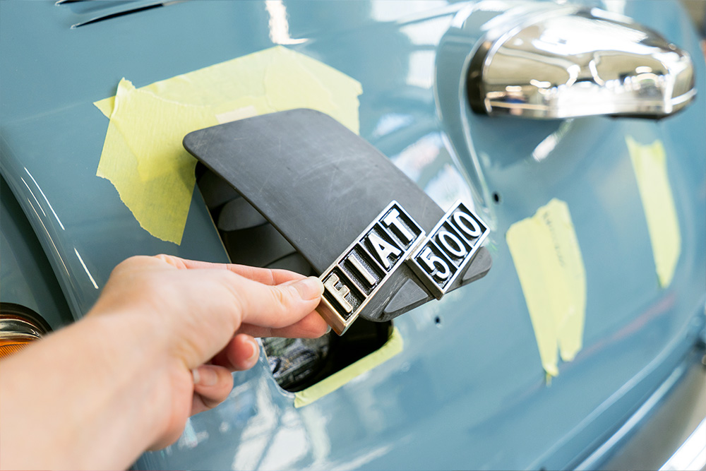 DSC09132_manufakturmarton_webgallerie Eine Öffnung am Heck des blau lackierten Fiat 500, die mit einer Klappe aus Kunststoff verschlossen werden kann. Im Bild ist diese halb geöffnet, an ihr ist das Logo "Fiat 500" angebracht.