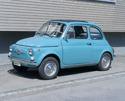 DSC00558_manufakturmarton_webgallerie2 Ein mittelblauer Fiat Nuova 500 steht vor einem Gebäude mit Holzfassade. Das Fahrzeug ist von der Seite fotografiert.