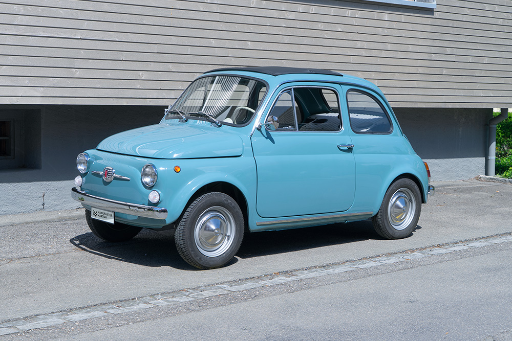 DSC00558_manufakturmarton_webgallerie2 Ein mittelblauer Fiat Nuova 500 steht vor einem Gebäude mit Holzfassade. Das Fahrzeug ist von der Seite fotografiert.